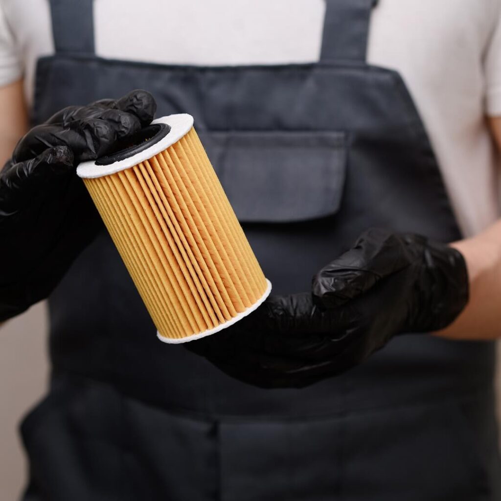 Mechanic wearing black gloves and a work apron holding a cylindrical hydraulic oil filter.
